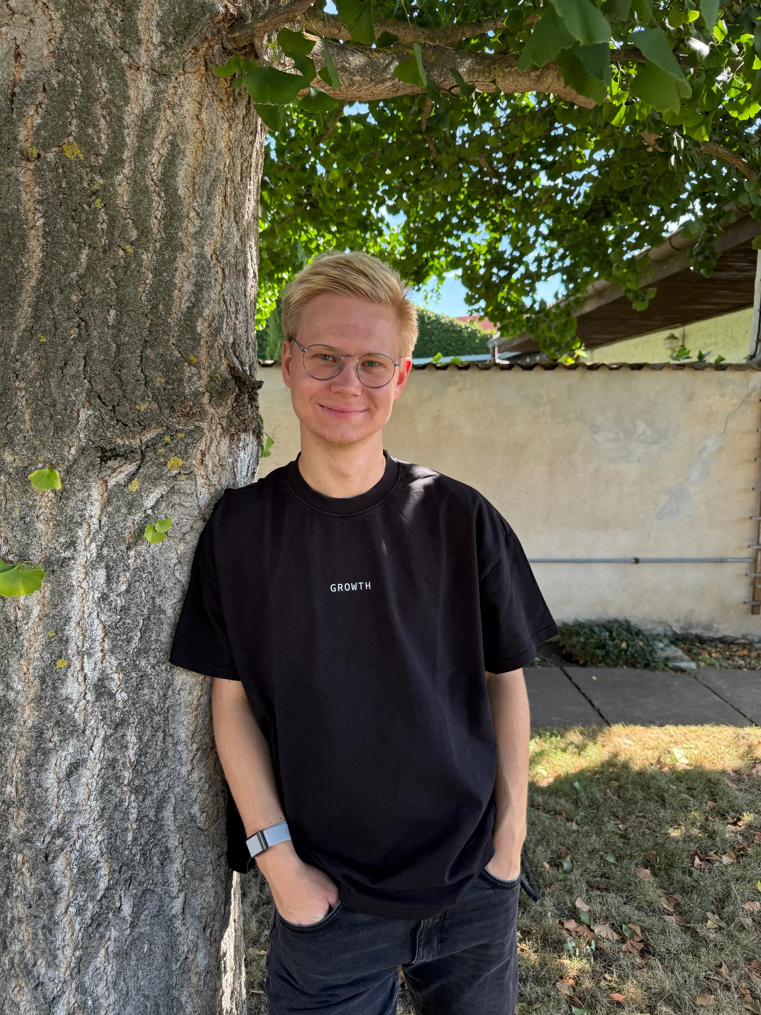 Erik Engler, lächelnd in schwarzem T-Shirt, entspannt an einen Baumstamm gelehnt.