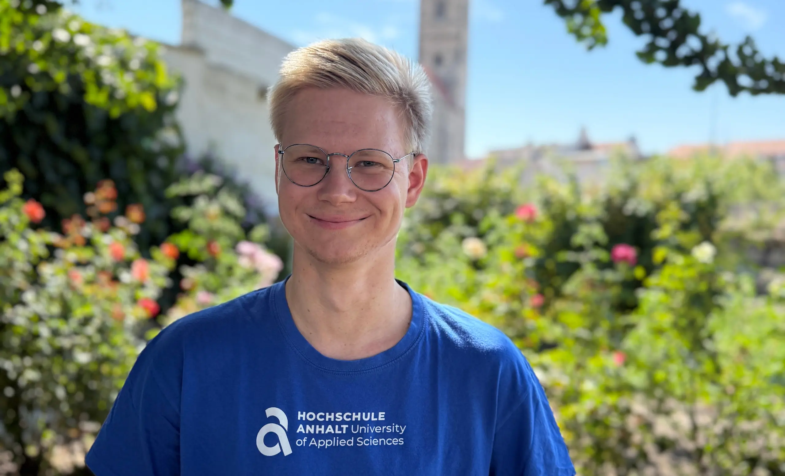 Erik Engler, lächelnd im blauen T-Shirt der Hochschule Anhalt; im Hintergrund unscharf ein Kirchturm und blühende Sträucher.