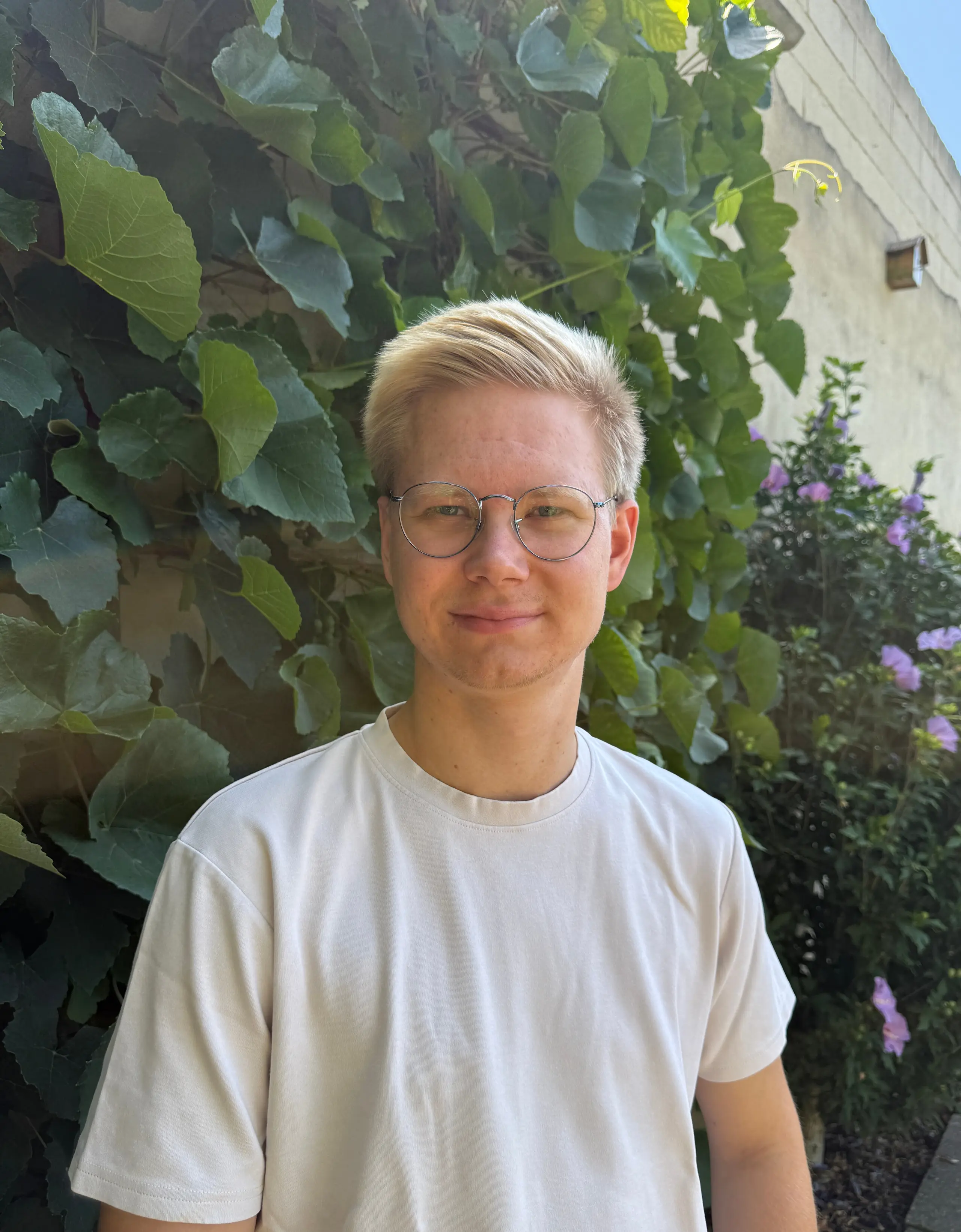 Erik Engler mit runder Brille, lächelnd in hellem T-Shirt vor einer dicht begrünten Pflanzenwand.